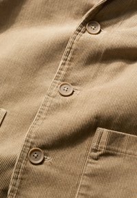 Gaiši brūns korduroja audums ar vertikālām svītrām, ar vairākām pogām un priekšējo kabatu. Faktūras virsma ar m�īkstu apdari.