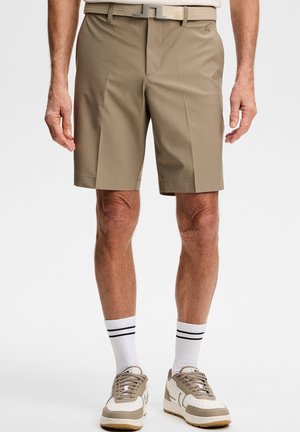 Mann trägt beige maßgeschneiderte Shorts, weiße Socken mit schwarzen Streifen und beige-weiße Sneakers, steht vor weißem Hintergrund.