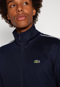 Marineblå zip-up jakke med høy krage. Har hvite striper langs skuldrene og et brodert grønt krokodillelogo på brystet.