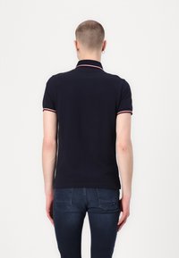 Polo shirt marine avec col et poignets rayés rouge et blanc, en coton. Il présente des manches courtes et un design ajusté, porté avec un jean foncé.