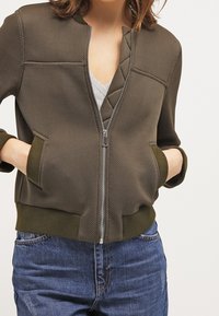 Oliwkowa kurtka zapinana na zamek, wykonana z teksturowanej tkaniny, z okrągłym dekoltem, dwiema przednimi kieszeniami i ściągaczami przy mankietach. Noszona na jasnoszarym topie.