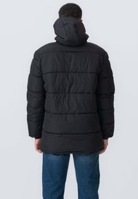 Sort pufferjakke med hætte, der har horisontale kviltede sektioner og elastiske manchetter, båret over blå jeans.