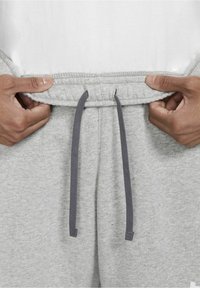 Pantalones de chándal grises con cinturilla elástica y cordón, que presentan una textura suave y un ajuste entallado. Manos sujetan la cinturilla para ajustarlos.