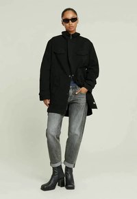 Μαύρο μπουφάν με φερμουάρ και δύο μπροστινές τσέπες, συνδυασμένο με γκρι τζιν με φθορά και ρολάρισμα στους ώμους, καθώς και μαύρες γόβες με χαμηλό τακούνι, φορεμένο με γυαλιά ηλίου.