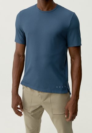 Mann, der ein eng anliegendes blaues kurzärmeliges Rippshirt und beigefarbene Hosen trägt, vor einem schlichten hellen Hintergrund stehend.