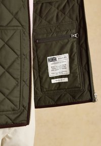 Gesteppte dunkelgrüne Jacke geöffnet, um eine innere Reißverschlusstasche und ein Etikett mit der UK-Flagge und dem Markentext „Joules“ zu zeigen.