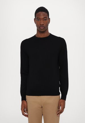 Maglione nero a maglia con scollo rotondo, maniche lunghe e una texture liscia, abbinato a pantaloni beige. Nessun motivo o accento visibile.