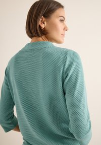 Camicia testurizzata color teal con un motivo a pois, vestibilità comoda e maniche a tre quarti. Presenta un colletto classico e un design con yoke posteriore.
