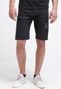 Svarta denimshorts med slitna revor, vanlig passform och uppvikt fåll. Kombinerat med ljusgrå sneakers på en neutral bakgrund.