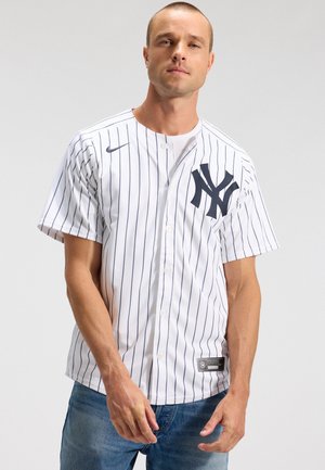 Mann trägt ein weißes New York Yankees Baseball-Trikot mit Nadelstreifen und blaue Jeans, steht vor einem einfarbigen hellen Hintergrund.