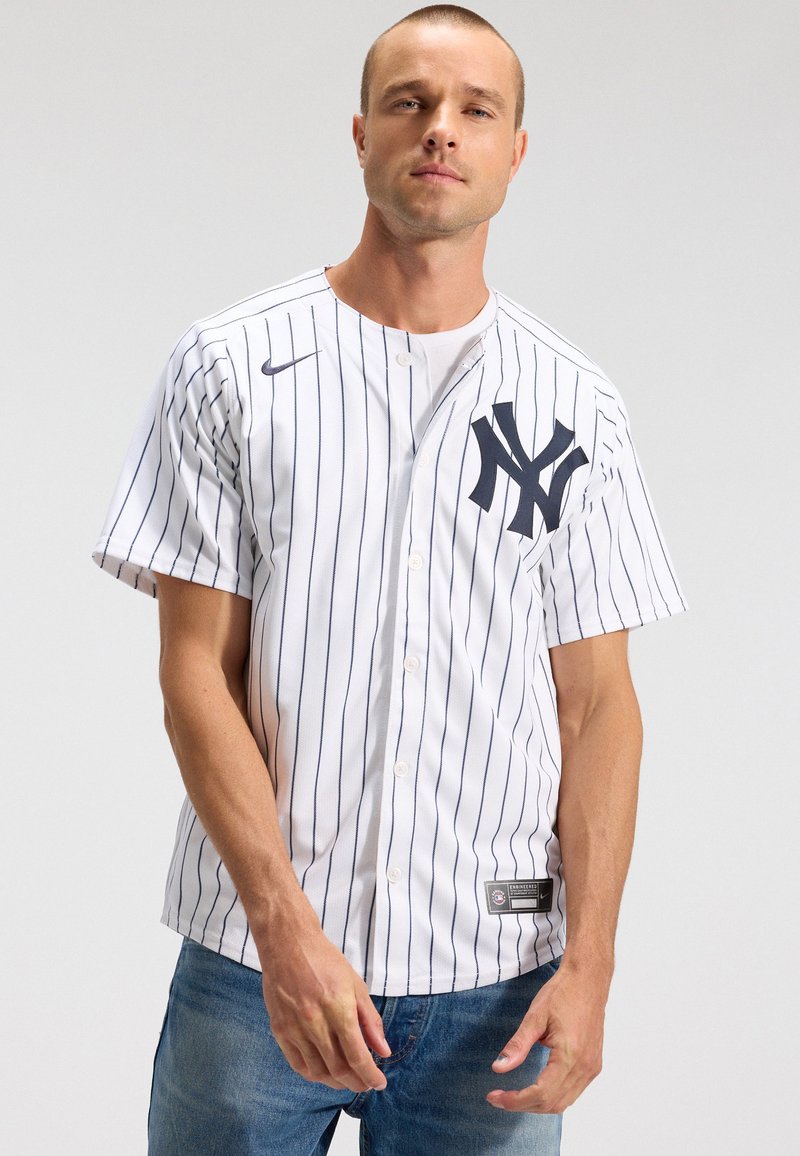 Άνδρας που φοράει λευκό μπέιζμπολ τζέρσεϊ New York Yankees με ρίγες και μπλε τζιν, στέκεται μπροστά σε απλό φωτεινό φόντο.
