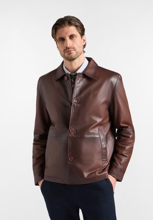 Uomo che indossa una giacca di pelle marrone scuro sopra una camicia con colletto e cravatta, in piedi con le mani nelle tasche davanti a uno sfondo semplice.