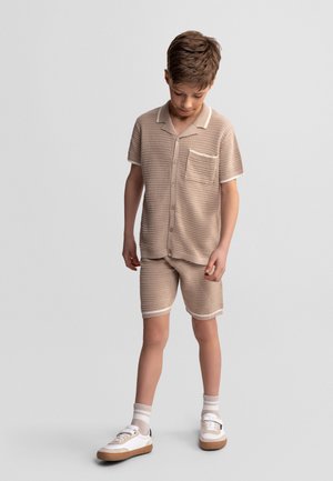 Jongen staand, draagt een beige gebreid overhemd met knopen en bijpassende korte broek, witte sokken en witte sneakers, kijkt naar beneden.