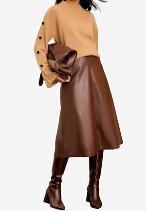 Femme portant un pull en maille camel avec des boutons noirs sur les manches, une jupe mi-longue en cuir marron, des bottes marron montantes jusqu'aux genoux, tenant un sac marron.