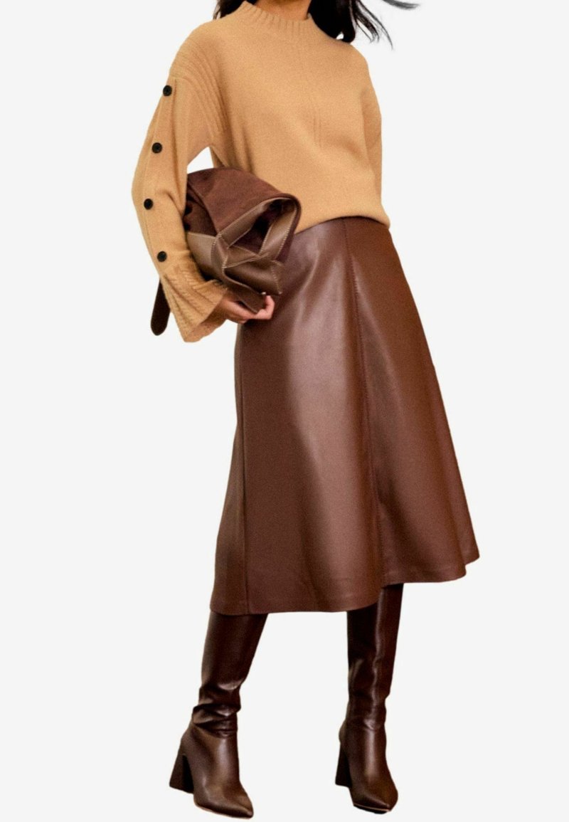 Femme portant un pull en maille camel avec des boutons noirs sur les manches, une jupe mi-longue en cuir marron, des bottes marron montantes jusqu'aux genoux, tenant un sac marron.