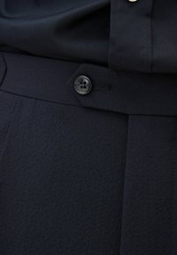 Pantaloni blu scuro con una texture liscia, un design semplice, chiusura con bottone e pieghe sul davanti per un aspetto su misura.