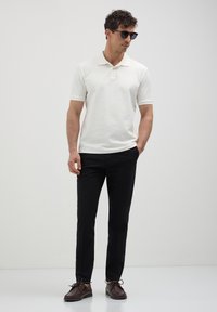 Polo shirt crème à manches courtes, pantalon noir ajusté, et chaussures à lacets marron, complétés par des lunettes de soleil noires. Design épuré et moderne.