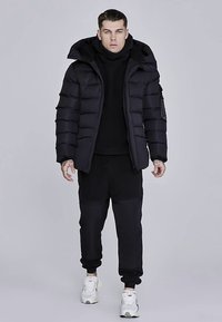 Črna puhovka z ruto s kapuco, s horizontalnim prešitim vzorcem in zadrgo po celotni dolžini, nošena preko črnega puloverja in trenirke.