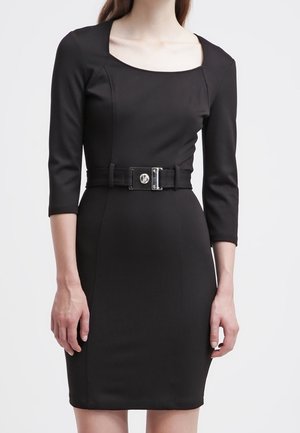 Frau trägt ein eng anliegendes schwarzes Kleid mit Dreiviertelärmeln und einem breiten Gürtel mit einer quadratischen silbernen Schnalle an der Taille.