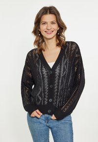 Fekete kötött kardigán, laza szabással, V-dekoltázzsal, mintás nyitott szövettel és hat fekete gombbal. Kék farmernadrággal párosítva.