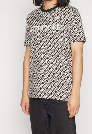 Homme portant un T-shirt à motif géométrique noir et blanc avec le texte "TOMMY HILFIGER" sur la poitrine et un pantalon noir.
