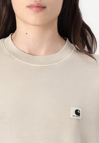 Felpa beige con bordo a coste, caratterizzata da una patch quadrata sul lato sinistro in alto. Il tessuto ha una consistenza liscia e morbida.