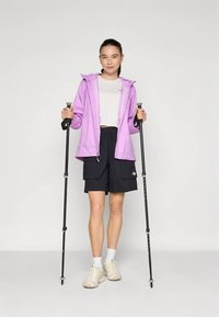 Νεαρή γυναίκα με μωβ σακάκι και μαύρες σορτς, κρατώντας μαύρα μπατόν πεζοπορίας, όρθια μπροστά σε απλό ανοιχτό φόντο.