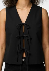 Juodas be rankovių topo su giliu V formos iškirpimu ir penkiais priekiais susietais raišteliais. Pagaminta iš lygaus audinio, su trumpu ilgiu ir paprastu dizainu.