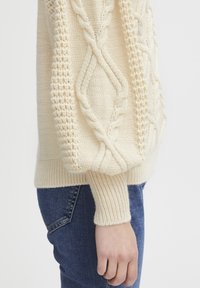 Pull en maille torsadée de couleur crème avec des motifs texturés sur les manches et une taille côtelée, associé à un jean en denim bleu.