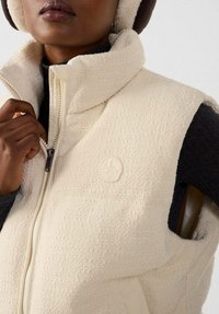 Beige Fleeceweste mit hohem Kragen, Reißverschluss und Markenpatch. Verfügt über eine strukturierte Oberfläche und kontrastierende schwarze Ärmelkante.