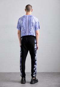 Gaiši zils rakstainais krekls ar īsām piedurknēm apvienots ar melniem sporta biksēm, kurām sānos ir zili ziedu motīvi, un melnām sporta apavām.