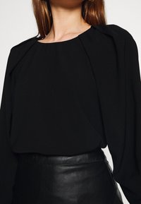 Blouse noire avec un col rond, des épaules plissées et des manches longues. Associée à une jupe en cuir taille haute pour un look monochrome.