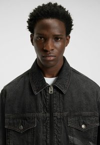 Zwarte denimjack met rode stiksels, ritsluiting en twee voorzakken. Klassiek kraagontwerp met getextureerde stof.