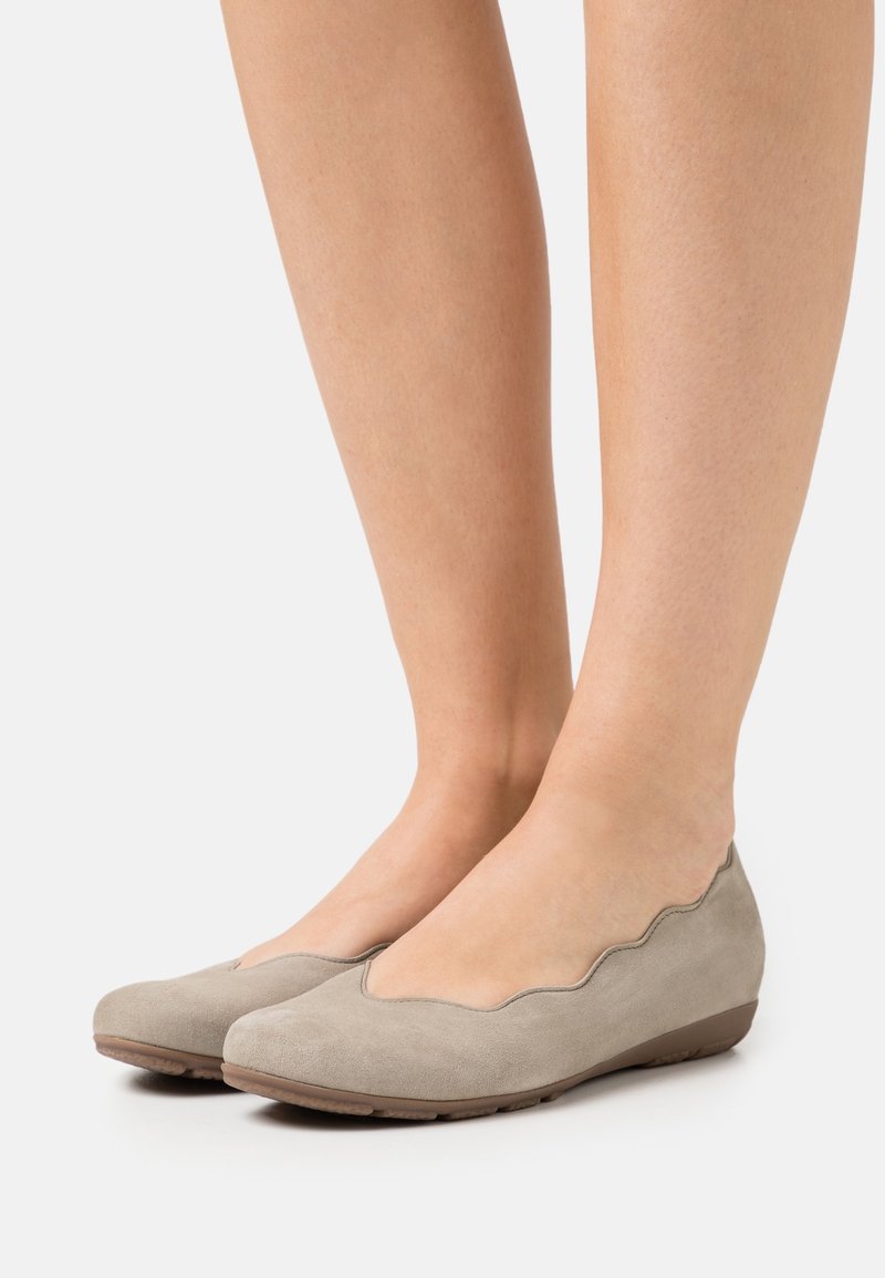 Pieds chaussés de ballerines en daim beige clair avec un bord festonné et un petit talon compensé sur fond blanc.