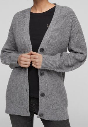 Personne portant un cardigan en maille gris avec des boutons noirs sur une chemise noire et un pantalon sombre, mains tenant les bords avant du cardigan.