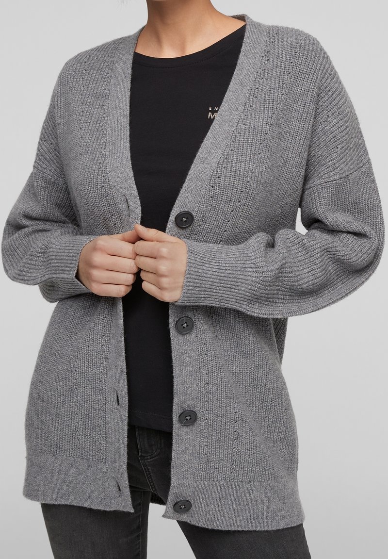 Personne portant un cardigan en maille gris avec des boutons noirs sur une chemise noire et un pantalon sombre, mains tenant les bords avant du cardigan.