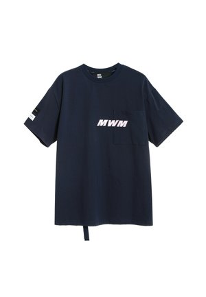 Hombre de espaldas vistiendo una camiseta azul marino con diseño paisley rosa y pantalones cortos beige, calcetines blancos y zapatos gris claros.