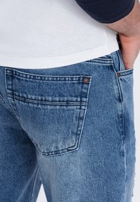 Jeans de mezclilla azul desvanecidos con un detalle en el bolsillo trasero y acentos de costura. La tela parece suave con una ligera variación de textura.