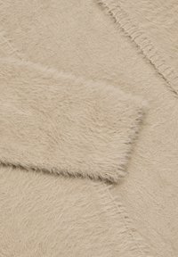 Beige tekstiil faux karvadega, millel on pehme ja karvane tekstuur. Kangas näitab veidi ebaühtlast pinda, mis esitleb erinevaid karvade pikkusi.