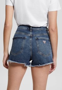 Denimshorts med højtaljet design, blå farve, frynset kant og baglommer med en lille logo-patch. Glat tekstur, afslappet design.