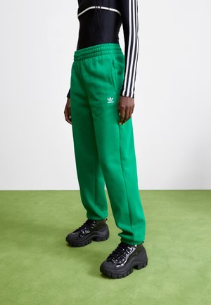 Personne portant un pantalon de survêtement Adidas vert vif et des baskets noires épaisses, debout sur un sol vert clair devant un fond blanc.
