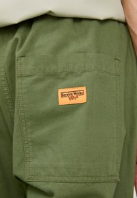 Pantalons cargo vert olive avec une grande poche arrière arborant une étiquette rectangulaire orange "Service Works". Fabriqués en tissu robuste avec des coutures visibles.