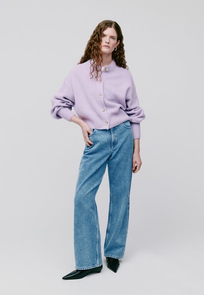 Lavendelfarbene Strickjacke mit großen Knöpfen, kombiniert mit hoch geschnittenen, weit geschnittenen blauen Jeans und spitzen schwarzen Schuhen.