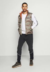 Mies seisoo pukeutuneena kamouflagepufferi-liiviin, valkoiseen pitkähihaiseen paitaan, mustiin juoksuhousuihin ja ruskeisiin vaelluskengiin, taustalla yksinkertainen tausta.