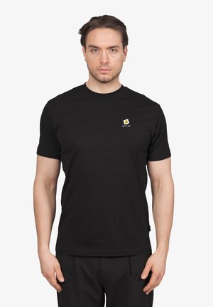 Mann trägt ein schwarzes T-Shirt mit einer kleinen Grafik eines Spiegeleis und dem Text "eggsitire safe" auf der linken Brust, stehend vor einem einfarbigen Hintergrund.