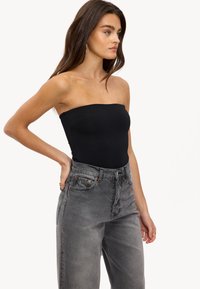 Femme aux longs cheveux bruns, portant un top noir sans bretelles et un jean gris délavé taille haute, se tenant avec une main sur la hanche, regardant sur le côté.