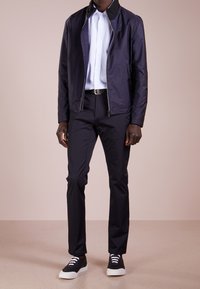 Donkerblauwe lichtgewicht jas met een zwarte kraag, gecombineerd met een lichtblauwe overhemd, geklede broek en zwarte sneakers met witte veters.