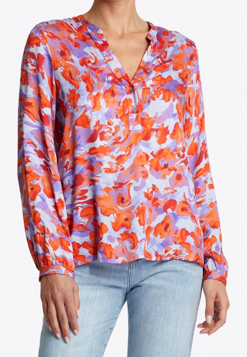 Femme portant un chemisier à manches longues avec un motif floral abstrait orange et lavande et un jean bleu clair.
