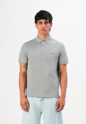 Mann med kort svart hår iført en grå polo-shirt og lyseblå jeans, stående mot en hvit bakgrunn.