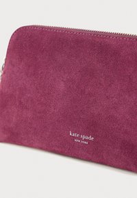 Fioletowa kopertówka zamszowa z zamkiem błyskawicznym. Na przodzie znajduje się wytłoczone logo "kate spade NEW YORK" w białym kolorze. Gładka faktura.